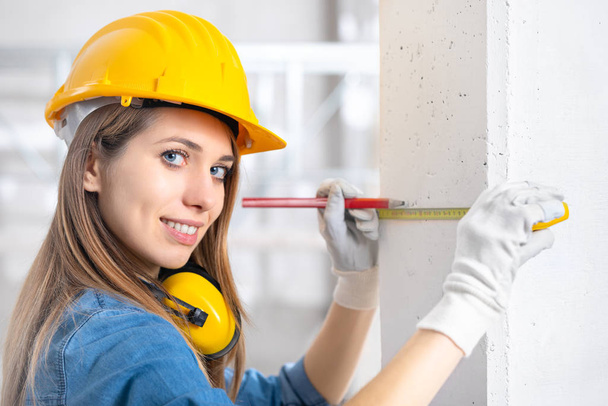 Young female construction worker in yellow hardhat and gloves measuring the wall with measuring tape and turning to smile at camera - 写真・画像