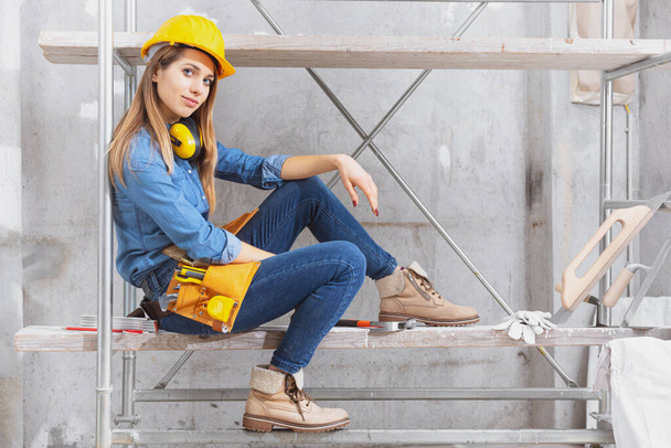 Trabalhadora da construção jovem capaz de usar um cinto de ferramentas e um chapéu duro sentado no andaime interior em uma sala em construção
 - Foto, Imagem