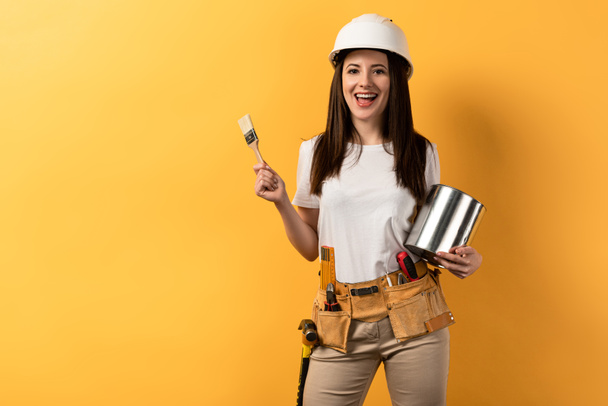 smiling handywoman holding paint brush and paint can on yellow background  - Foto, imagen
