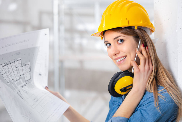 Mladá žena inženýr nebo architekt drží plán v ruce, jak si povídá na svém smartphonu na místě v místnosti ve výstavbě otáčí k úsměvu na kameru - Fotografie, Obrázek