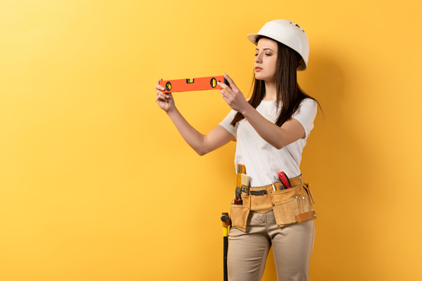 concentrated handywoman in helmet holding spirit level on yellow background  - Photo, Image