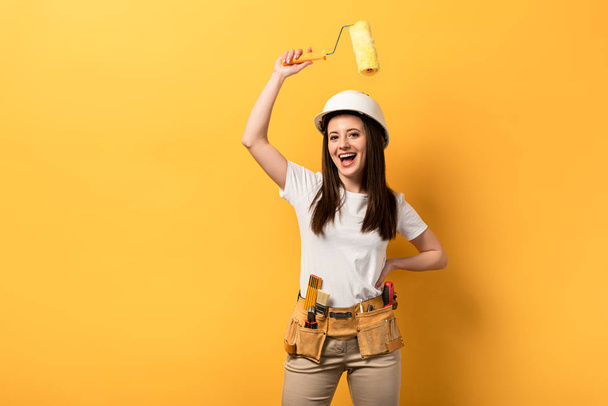 lachende handywoman met hand op heup holding paint roller op gele achtergrond  - Foto, afbeelding