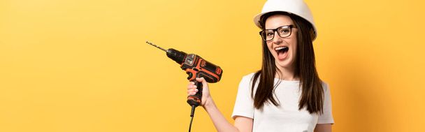 panoramic shot of positive handywoman screaming and holding drill on yellow background  - Photo, Image