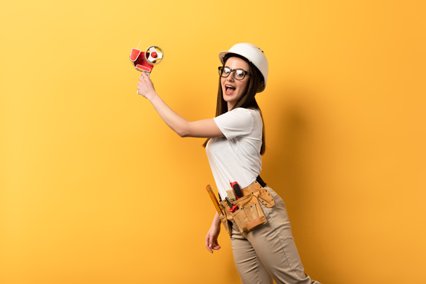šokované údržbářka drží pásku dávkovač na žlutém pozadí s kopírovacím prostorem  - Fotografie, Obrázek