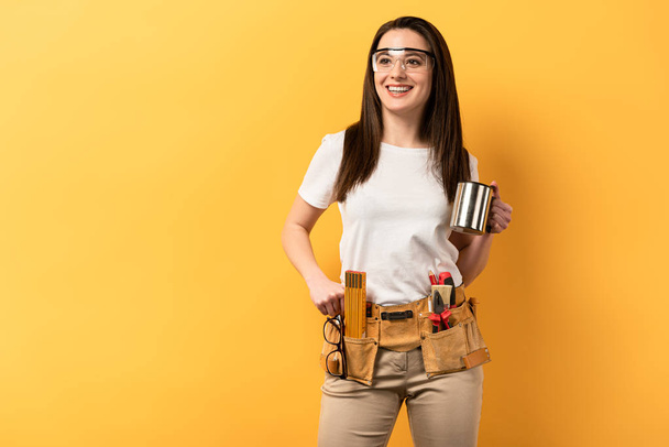 smiling handywoman holding metal cup on yellow background  - Photo, Image