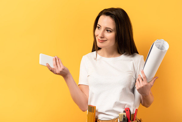 smiling handywoman holding smartphone and blueprint on yellow background  - Foto, Imagen
