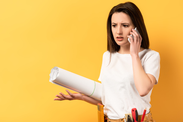 shocked handywoman talking on smartphone and holding project on yellow background  - Photo, Image