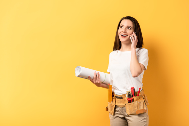 smiling handywoman holding smartphone and blueprint on yellow background  - Foto, Imagen