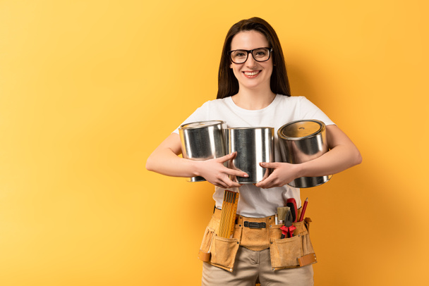 smiling repairwoman holding paint cans and looking at camera on yellow background  - Photo, Image