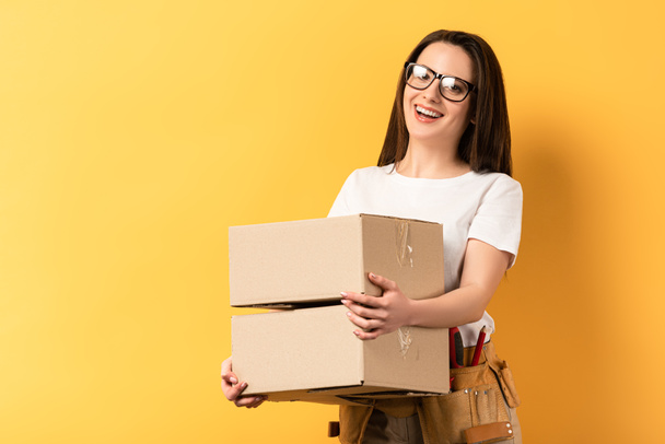 smiling repairwoman holding boxes and looking at camera on yellow background  - Photo, Image