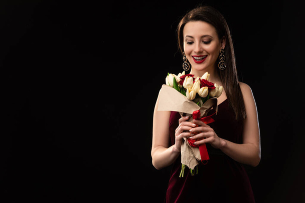 excited woman in dress looking at bouquet isolated on black - Fotografie, Obrázek
