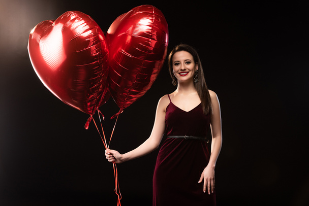 smiling woman in dress holding heart-shaped balloons in 14 february on black background  - Photo, Image