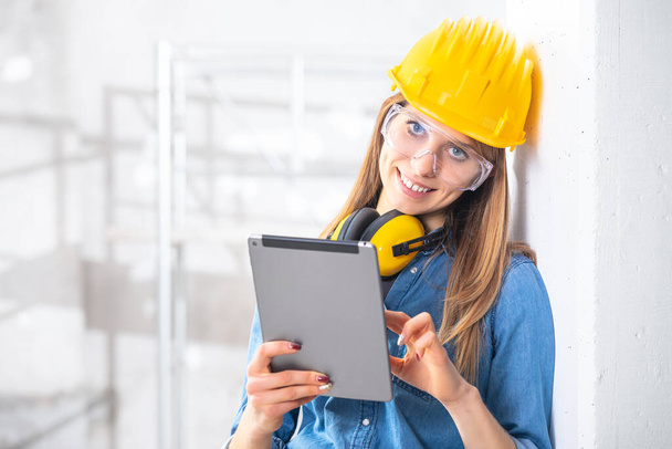 Mosolyogva fiatal női építőmunkás visel keménykalap és fülvédő segítségével tabletta számítógép a helyszínen, magas kulccsal háttér - Fotó, kép