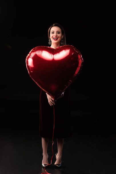 smiling woman in dress holding heart-shaped balloon on black background  - Fotó, kép