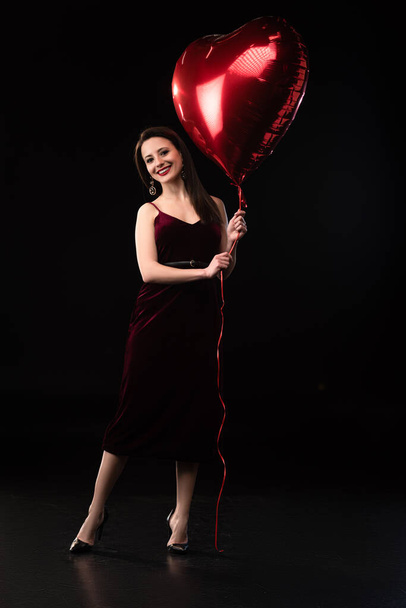 smiling woman in dress holding heart-shaped balloon on black background  - Фото, зображення