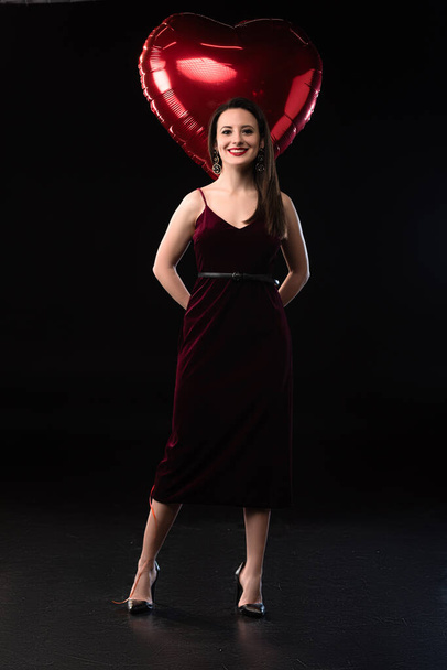 smiling woman in dress holding heart-shaped balloon on black background  - Foto, Imagem