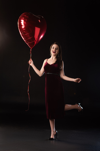 smiling woman in dress holding heart-shaped balloon on black background  - Foto, imagen
