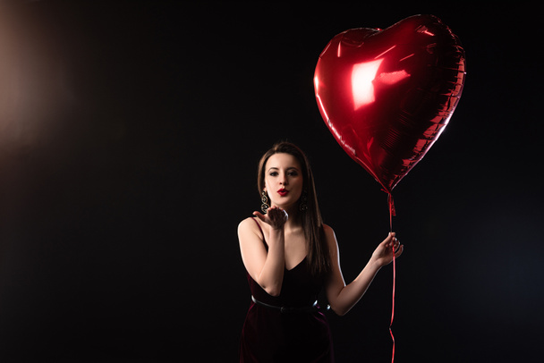 woman in dress holding heart-shaped balloon and blowing air kiss on black background  - Foto, imagen