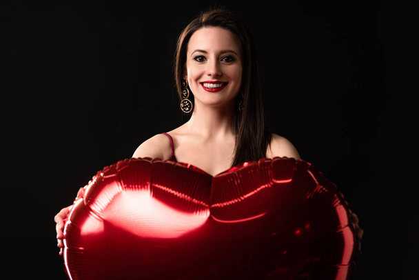 smiling woman holding heart-shaped balloon in 14 february isolated on black - Φωτογραφία, εικόνα
