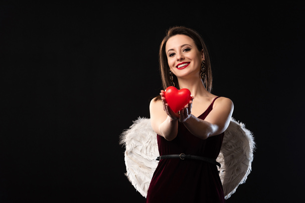 glimlachende vrouw in jurk met vleugels met hartvormig model in 14 februari geïsoleerd op zwart - Foto, afbeelding