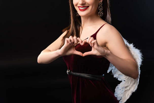 cropped view of smiling woman in dress with wings showing heart gesture isolated on black - Fotografie, Obrázek