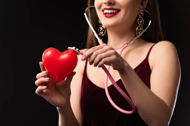 обрезанный вид улыбающейся женщины в платье, держащей модель в форме сердца и стетоскоп, изолированный на черном
 - Фото, изображение