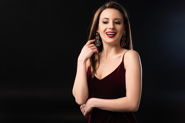glimlachende vrouw in jurk met oorbellen weg te kijken op zwarte achtergrond  - Foto, afbeelding