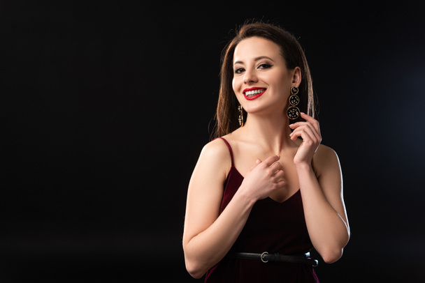 smiling woman in dress with earrings looking at camera on black background  - Foto, Bild