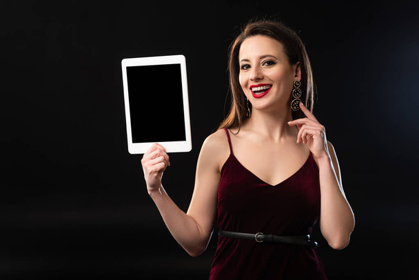 glimlachende vrouw in jurk houden digitale tablet op zwarte achtergrond  - Foto, afbeelding