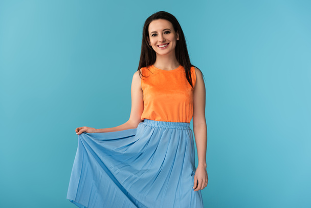 smiling and attractive woman looking at camera and holding skirt on blue background  - Фото, изображение