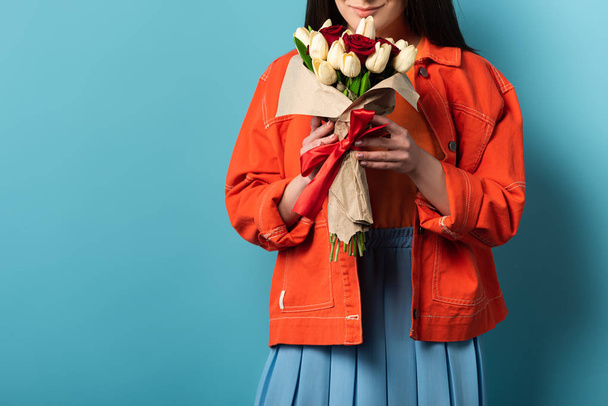 cropped view of smiling woman in jacket smelling bouquet on blue background  - Fotografie, Obrázek