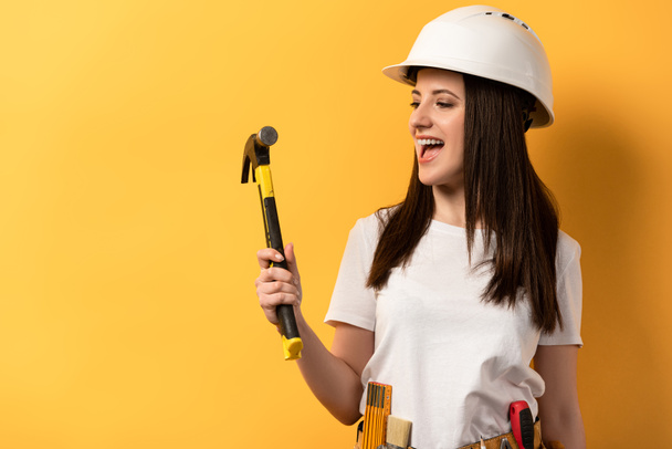 smiling handywoman in helmet holding hammer on yellow background  - Photo, image