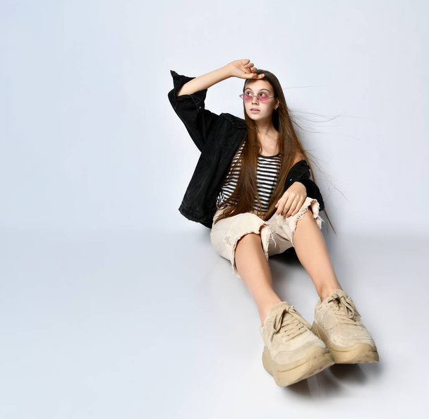 Teenage girl in striped t-shirt, black denim jacket, pants and sneakers. She is sitting on the floor isolated on white. Close up - Fotoğraf, Görsel