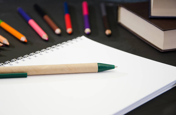 Onderwijsconcept. Notitieboekje met pen op de achtergrond van kleurpotloden en boeken op een zwarte tafel. Close-up. - Foto, afbeelding