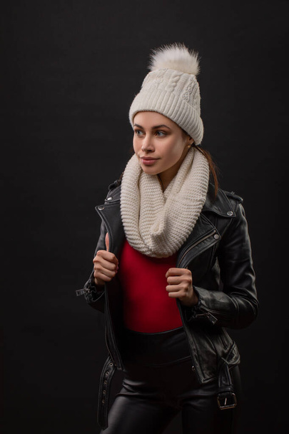 A stylish young woman in a black coat and knitted hat and snud poses on a black background. Autumn and winter women's fashion. - Фото, изображение