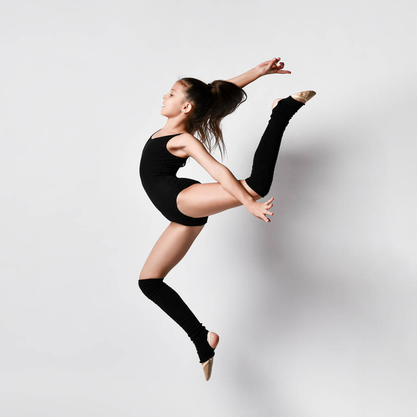 Teenage brunette girl gymnast with pigtail, in black leotard, knee socks and ballet shoes, performing exercises, posing isolated on white. Close-up. - Foto, Bild