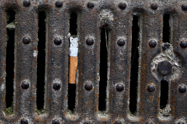 Cigarette in a rusty sewer grid, Dropped cigarette butt in canal, city environment. - Photo, Image