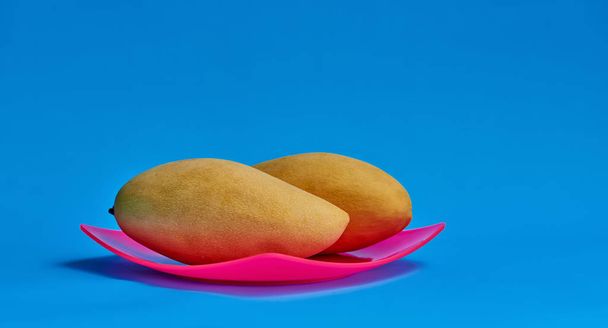 Two yellow mangoes like yin and yang on a pink plate. Blue background. Free space for text. - Φωτογραφία, εικόνα