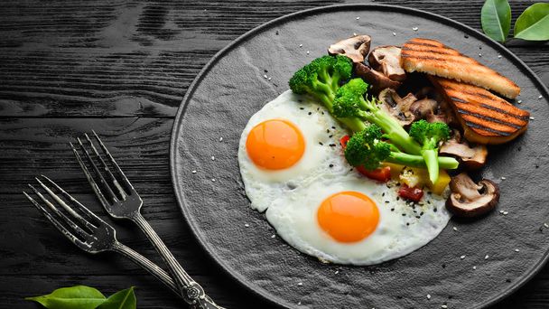 Healthy breakfast. Fried egg with broccoli, mushrooms and bread. On a black stone plate. Top view. - 写真・画像