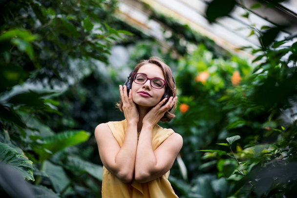 Young woman with headphones standing in greenhouse in botanical garden. - Foto, afbeelding