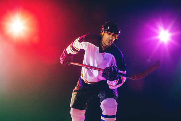 Male hockey player with the stick on ice court and dark neon colored background - Foto, afbeelding