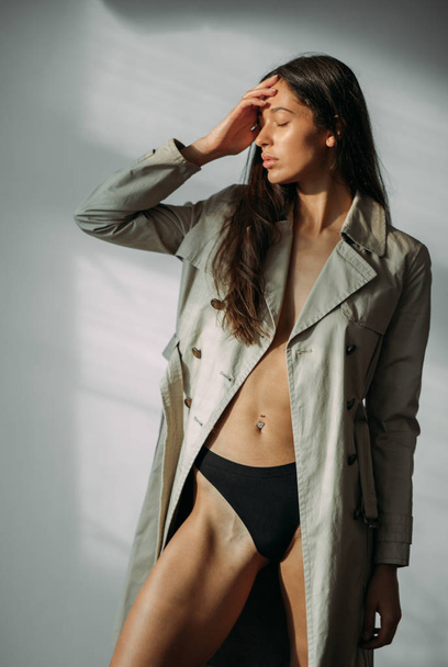Young sad woman in a coat and black bikini posing in the studio on a gray background. - 写真・画像