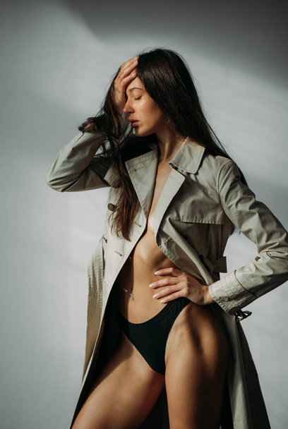 Young sad woman in a coat and black bikini posing in the studio on a gray background. - Foto, afbeelding