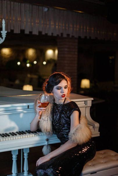 Portret van feestelijke schoonheid uit de jaren '20 in een restaurant. Jonge mooie vrouw in art-deco stijl, in zwarte jurk in een luxe interere Chicago 20s, het tijdperk van gangsters. Zanger aan de witte piano - Foto, afbeelding