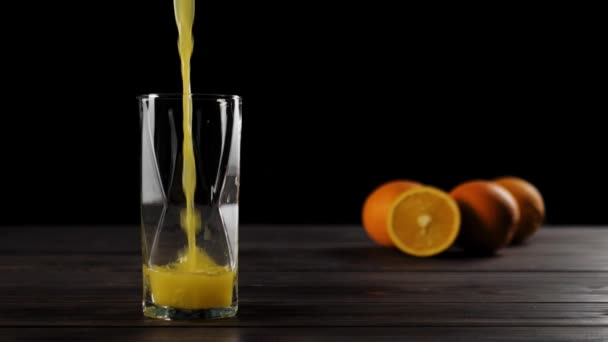 Große Aufnahme von frischem Orangensaft, der in Cassiopea-Glas gegossen wird, auf einem Holztisch mit köstlichen Orangen im weichen Fokus im Hintergrund vor schwarzem Hintergrund - Filmmaterial, Video
