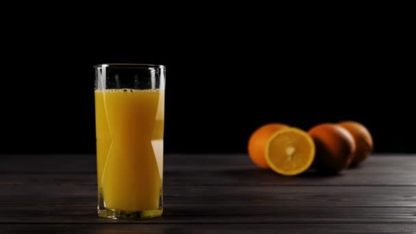 Широкий снимок кубиков льда, падающих в бокале Cassiopea со свежим апельсиновым соком, создающих спайсы и пузырьки на деревянном столе, украшенном апельсинами в мягком фокусе на заднем плане - Кадры, видео