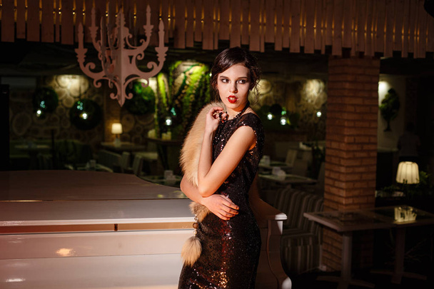Portrait of 20s style festive beauty in a restaurant. Young beautiful woman in art-deco style, in black dress in a luxurious interere Chicago 20s, the era of gangsters. Singer at the white piano - Photo, Image