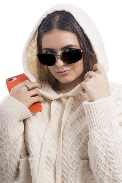 Fashionable girl in a white hoodie and sunglasses with smart phone in her hand on a white background. - Foto, Imagem