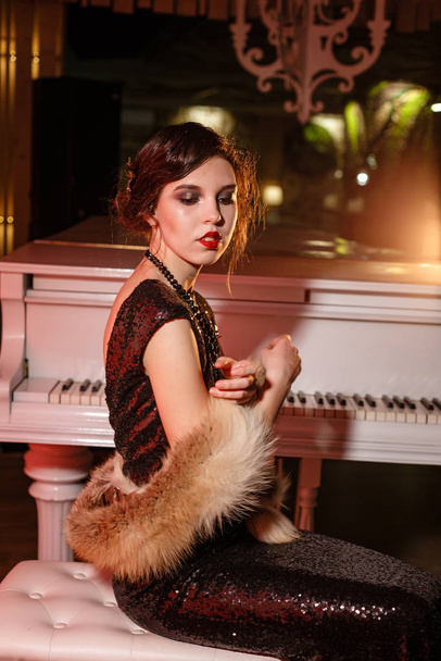 Portrait of 20s style festive beauty in a restaurant. Young beautiful woman in art-deco style, in black dress in a luxurious interere Chicago 20s, the era of gangsters. Singer at the white piano - Photo, Image