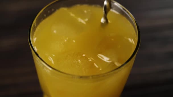 Widok z góry łyżka barmana mieszając kostki lodu ze świeżym sokiem pomarańczowym w wysokiej szklance przeciwko drewnianym stole - Materiał filmowy, wideo